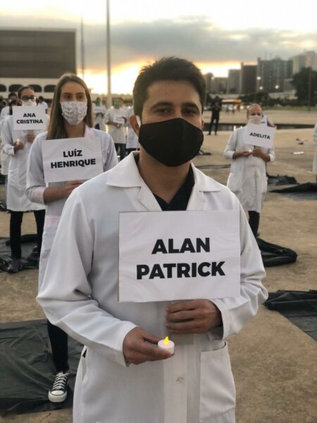 Pessoas com jaleco e máscara seguram placas com nome de mortos na pandemia e vela