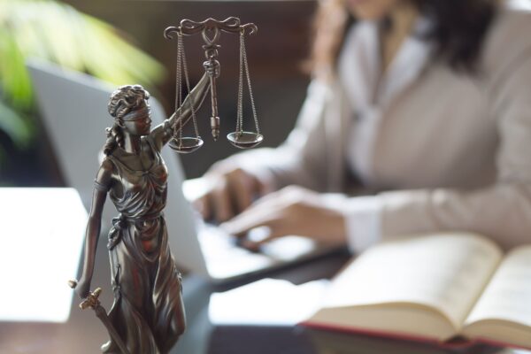 A imagem mostra uma estátua da deusa da justiça, segurando a balança, em destaque no primeiro plano. Ao fundo, de maneira desfocada, uma pessoa usando roupa formal trabalha em um laptop, com um livro aberto ao lado, simbolizando um ambiente de análise técnica ou jurídica. 