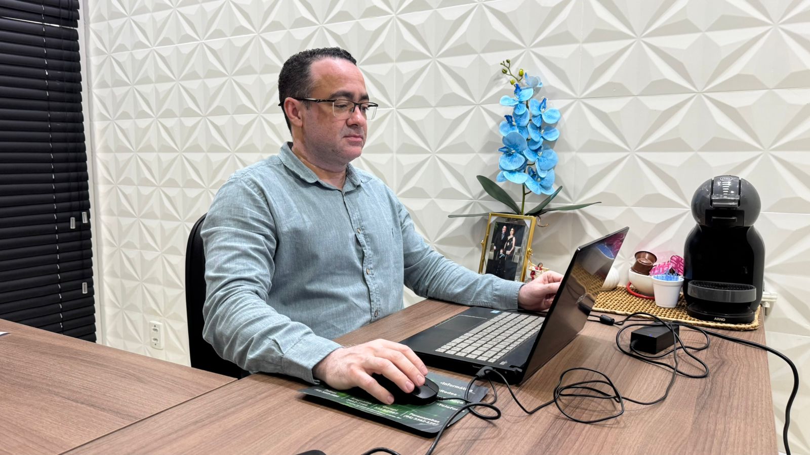 Homem branco, de óculos, sentado em uma mesa de escritório e olhando para o notebook