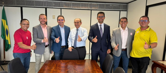 Contexto Geral: Uma fotografia colorida em plano médio de um grupo de sete homens adultos de diferentes idades e etnias, em pé, alinhados lado a lado, fazendo um gesto de 
