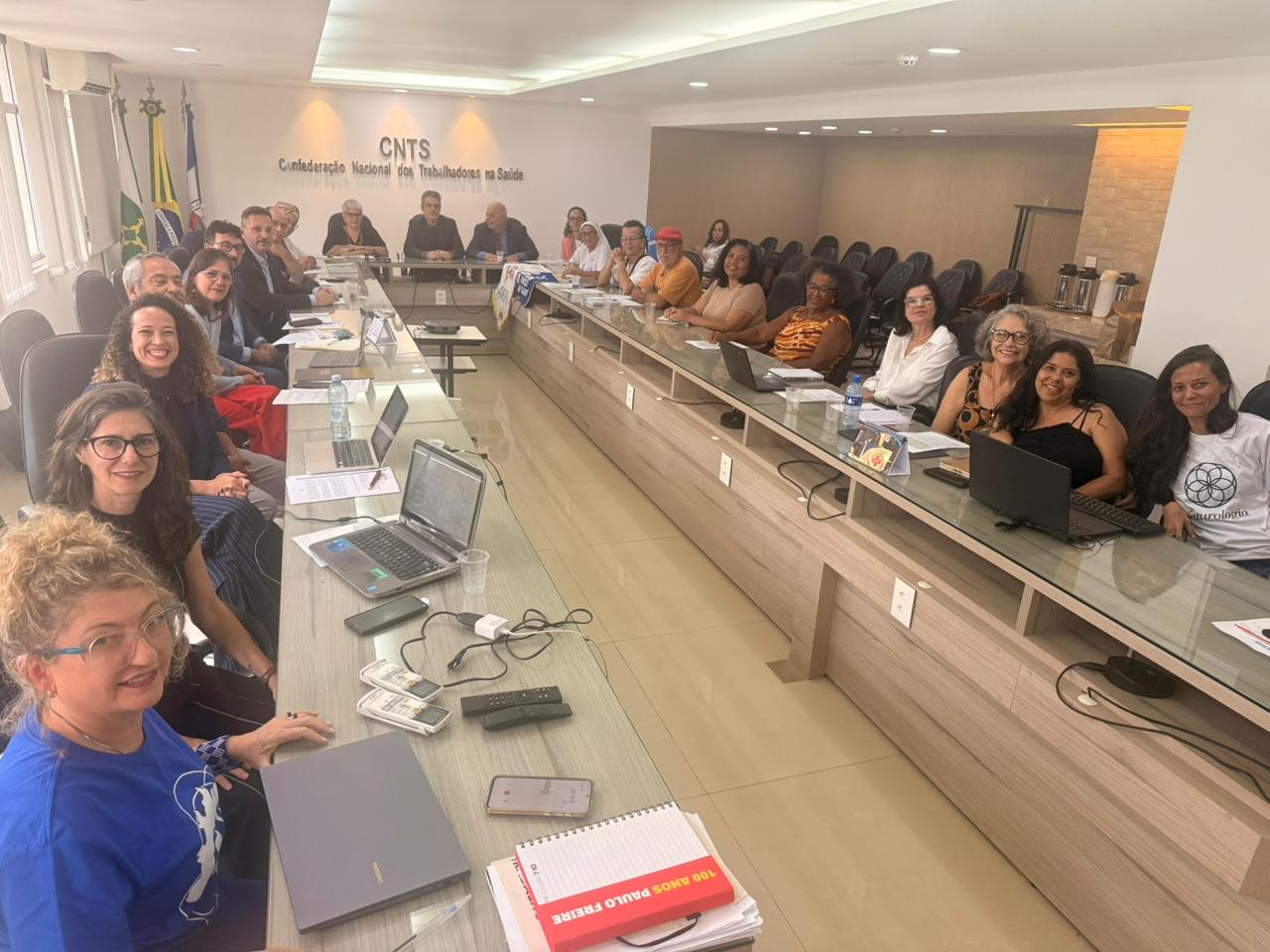 Reunião na sede da CNTS com cerca de 20 pessoas sentadas em uma grande mesa em formato de "U". Elas utilizam notebooks e documentos em uma sala ampla e iluminada. Ao fundo, o logotipo da entidade e as bandeiras oficiais.