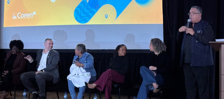 Paulo Murilo fala ao microfone em um palco durante evento do Coren-RJ. Ao lado dele, outras autoridades estão sentadas, acompanhando a programação do simpósio