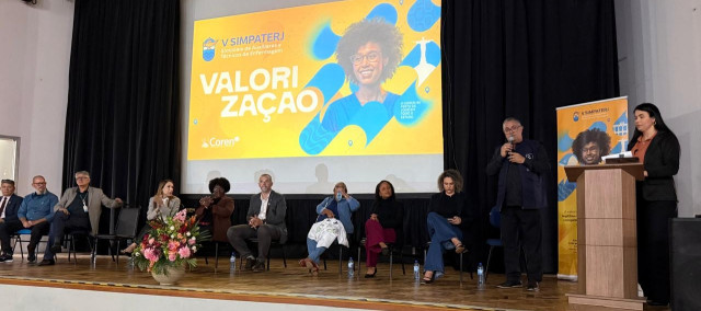 Paulo Murilo fala ao microfone em um palco durante evento do Coren-RJ. Ao lado dele, outras autoridades estão sentadas, acompanhando a programação do simpósio